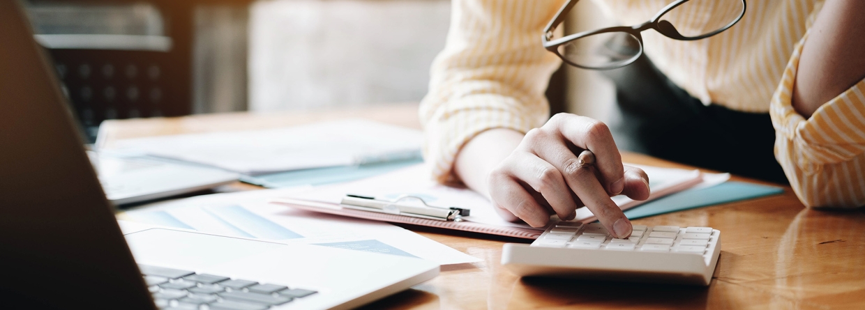 Vrouw met een bril in haar hand maakt berekeningen op een rekenmachine aan de hand van papieren op haar bureau