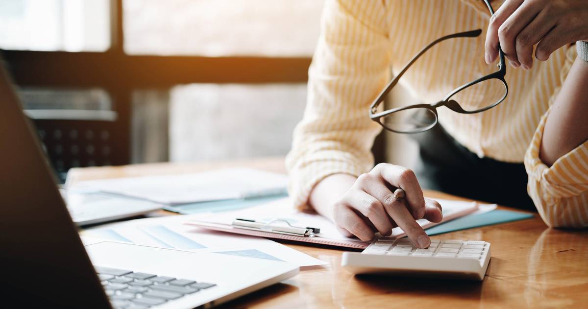 Vrouw met een bril in haar hand maakt berekeningen op een rekenmachine aan de hand van papieren op haar bureau